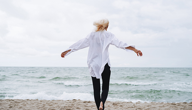 Frau am Strand von hinten zu sehen, mit ausgebreiteten Armen, blonde Haare im Wind, frei und gelöst barfuss Richtung Meer